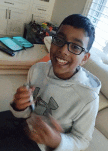 a young boy wearing glasses and a white under armour sweatshirt smiles for the camera