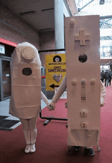 two people dressed as nintendo controllers holding hands in front of a sign that says comic games village