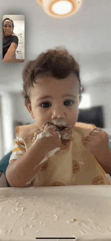 a baby wearing a bib is eating food while a woman looks on