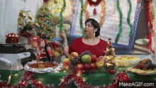 a woman in a red shirt is standing in front of a table filled with fruit and a christmas tree .