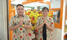 a man and a woman are posing for a picture in front of a tree with yellow flowers and a sign that says ta