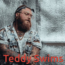 a man with a beard and tattoos is standing in front of a silver wall with the words teddy swims in red letters