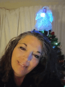 a woman smiles in front of a christmas tree with a lighted angel on top