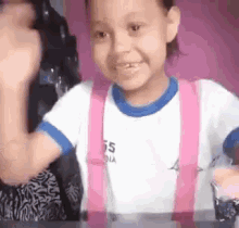 a little girl wearing pink suspenders and a white shirt is sitting at a table with a glass of milk .