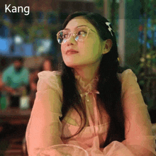 a woman wearing glasses sits at a table with kang written on the bottom right