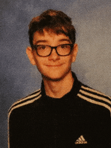 a young man wearing glasses and a black adidas shirt smiles for the camera