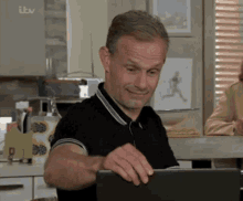 a man in a black shirt is using a laptop in a kitchen with itv written on the wall behind him