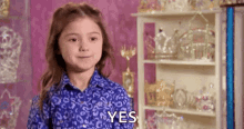 a little girl in a blue shirt is standing in front of a shelf with trophies and says `` yes '' .