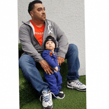 a man wearing a polo shirt sits on the grass holding a baby