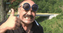 a man wearing sunglasses is giving a thumbs up in front of a sign that says cigoja