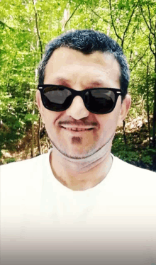 a man wearing sunglasses and a white shirt smiles in front of a forest