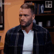 a man wearing a plaid shirt is standing in front of a bookshelf with the word sistas on the wall behind him