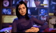 a woman in a purple turtleneck is sitting in front of a wall with pictures of faces on it .