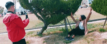 a man in a red jacket is taking a picture of a woman sitting under a tree in a park .