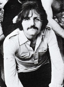 a man with long hair and a mustache is sitting in a black and white photo .