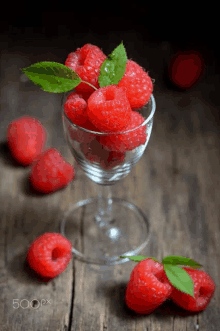 a glass filled with raspberries with leaves next to raspberries on a table