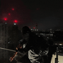 a man in a hoodie stands on a balcony at night with red lights in the background