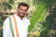 a man in a white shirt and orange and green sash smiles in front of a green leaf