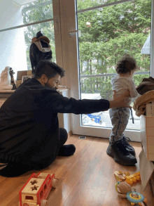 a man kneeling on the floor holding a baby while looking out a doorway