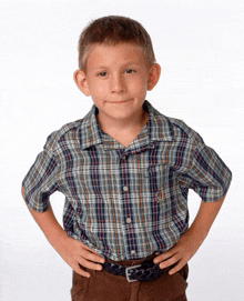 a young boy wearing a plaid shirt and brown pants stands with his hands on his hips