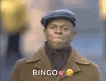 a man wearing a hat and a coat is standing in front of a building and says bingo .