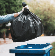 a person wearing white gloves is throwing a black garbage bag into a blue trash can