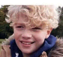 a young boy with curly blonde hair is smiling for the camera while wearing a fur hooded jacket .