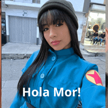 a woman wearing a blue jacket and a green beanie is standing in front of a sign that says hola mor