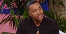 a young man wearing a black shirt and a necklace is smiling while sitting in front of a plant .