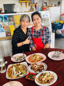 two women are sitting at a table with plates of food and one of them is wearing an apron that says ' thailand '