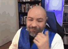 a bald man with a beard is giving a thumbs up while sitting in a chair in front of a bookshelf .