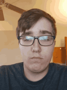 a young man wearing glasses looks at the camera with a ceiling fan behind him