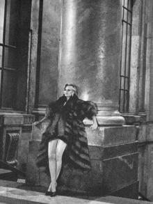 a black and white photo of a woman in a fur coat leaning against a pillar