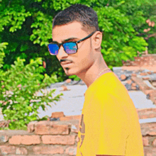 a young man wearing sunglasses and a yellow shirt stands in front of a brick wall