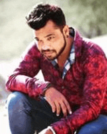 a man with a beard is sitting on a rock wearing a red shirt and blue jeans .