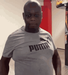 a bald man wearing a grey puma t-shirt is standing in a hallway .