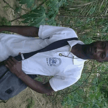 a man wearing a white shirt with a black strap around his neck is standing in front of a tree .