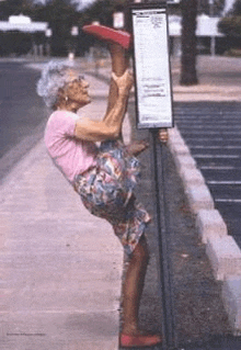 an elderly woman is doing a split while standing on a bus stop .