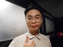 a young man wearing glasses and a white shirt gives a thumbs up