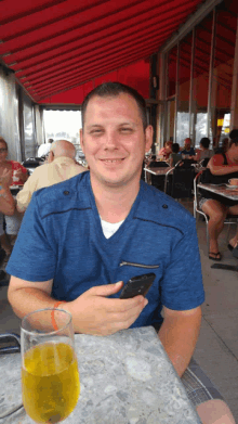 a man in a blue shirt is sitting at a table with a glass of beer and a cell phone