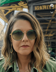 a woman wearing sunglasses and a green jacket stands in front of a building that says klatry 's