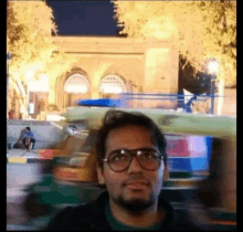 a man wearing glasses is standing in front of a blurry picture of a building at night .