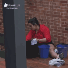 a woman in a red shirt is sitting on the floor with buckets and a playplus logo in the background