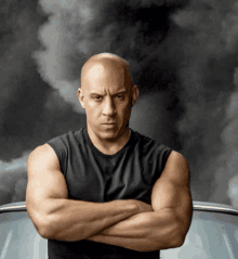 a bald man with his arms crossed is standing in front of a car