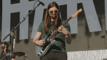 a man wearing sunglasses and a green shirt plays a guitar in front of a stage with a sign that says stagecoach