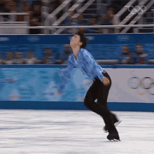 a man in a blue shirt and black pants is skating on a rink with the olympic rings in the background