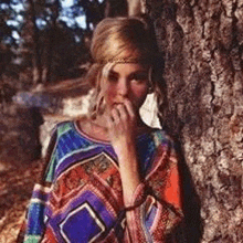 a woman in a colorful dress is standing next to a tree and covering her mouth with her hand .