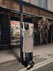 a man in a trench coat leans against a pole with his arms crossed in front of a building with a sign on it