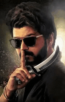 a man with a beard wearing sunglasses and a knife is holding his finger to his mouth .