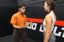 a woman in an orange shirt is talking to another woman in front of a wall with the letter l on it